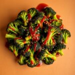 Close-up of Smashed Sambal Broccoli topped with a drizzle of spicy sambal sauce and garnished with sesame seeds on a warm-colored background, highlighting the vibrant green broccoli florets and rich, tangy glaze.