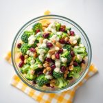 A bowl of Broccoli Crunch Salad featuring fresh broccoli, dried cranberries, and nuts, presented on a yellow checkered cloth.