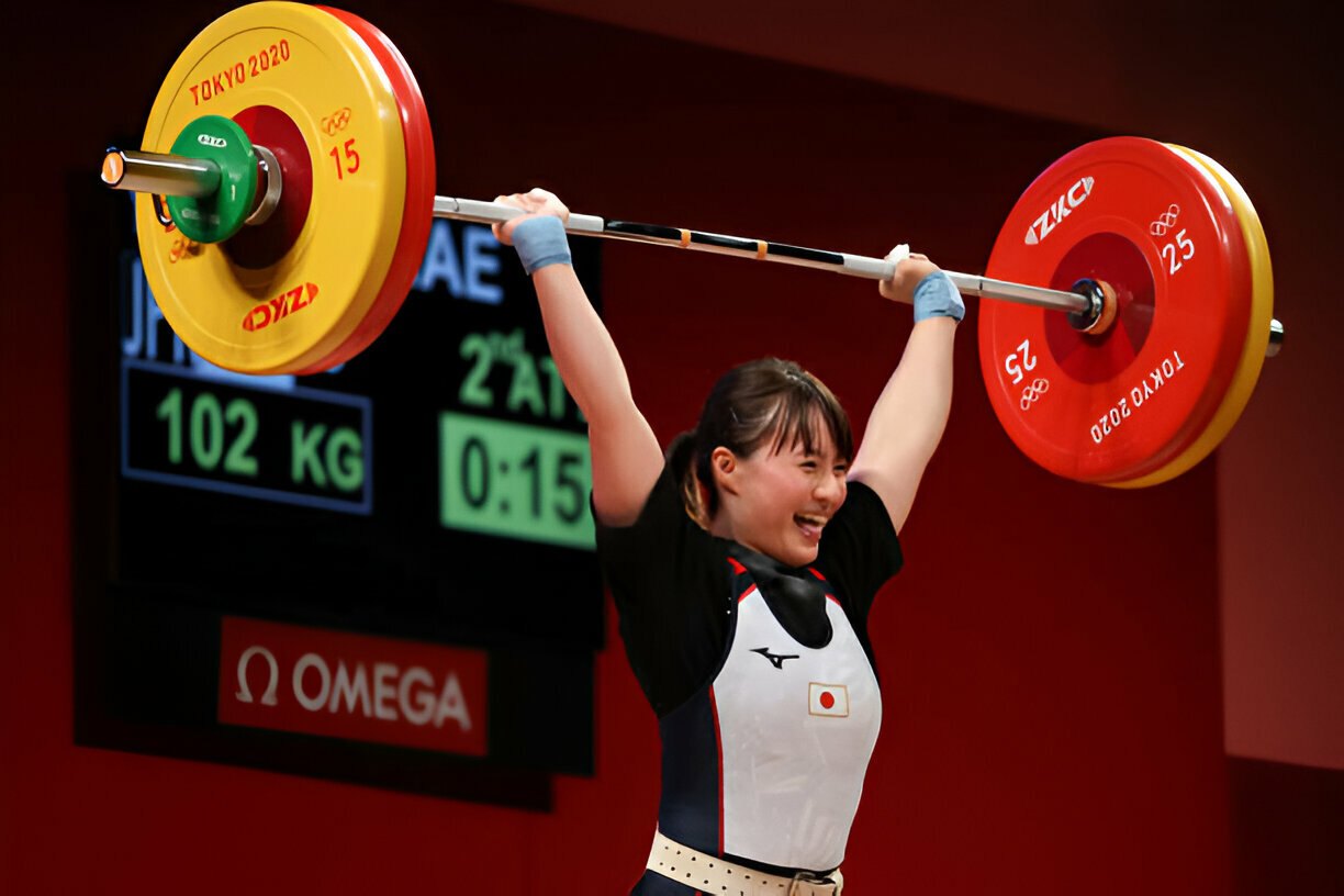 An athlete performing a successful lift in Olympic weightlifting at the Tokyo 2020 Olympics, smiling with joy as she holds the barbell overhead.