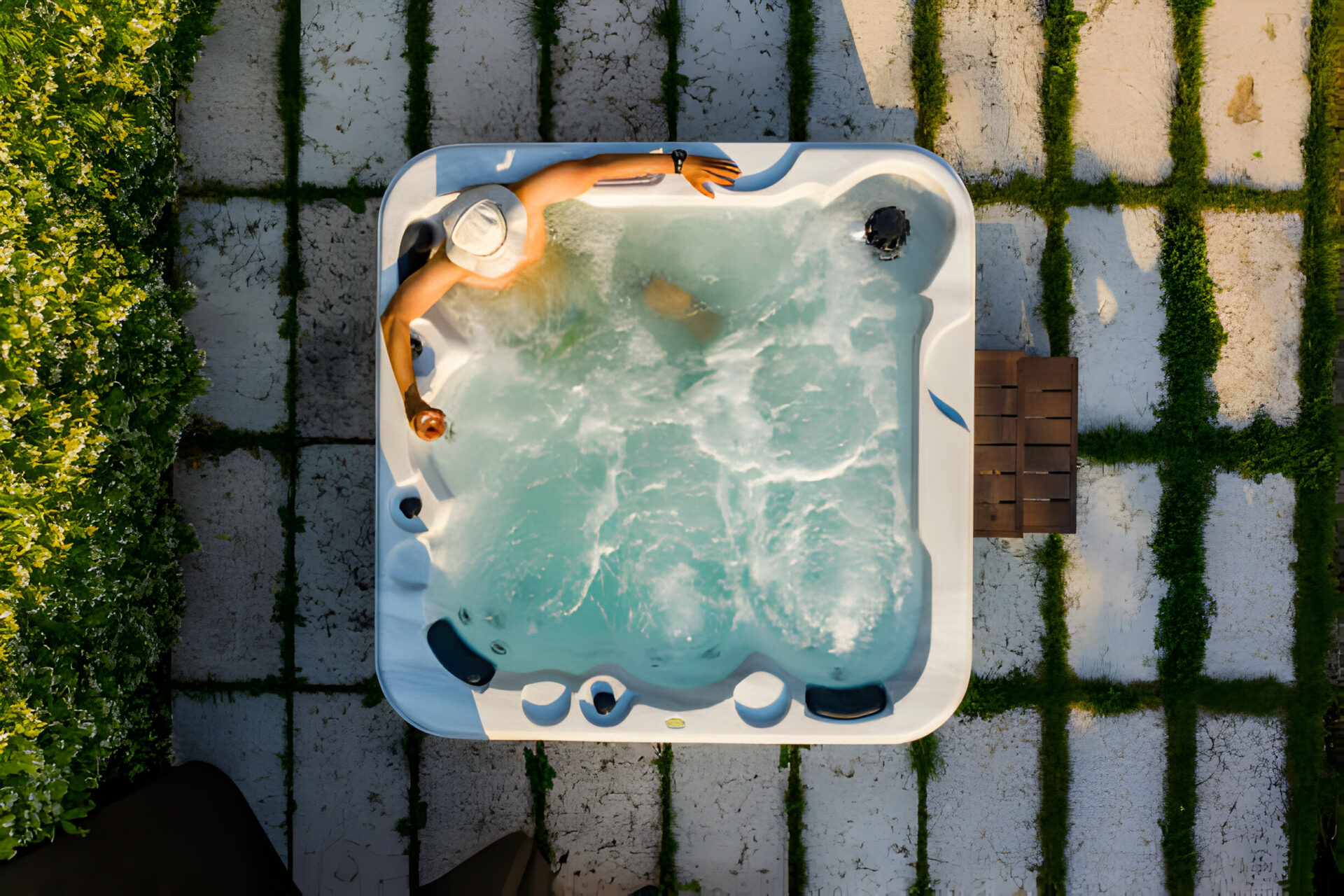 Top view of a person relaxing in a hot tub with a drink in hand, surrounded by a modern outdoor setting with greenery and paved stone tiles.